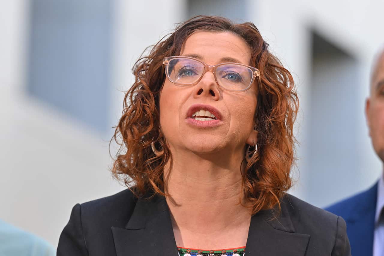 A woman with shoulder-length hair and glasses and wearing a black suit is captured as she talks to the cameras.