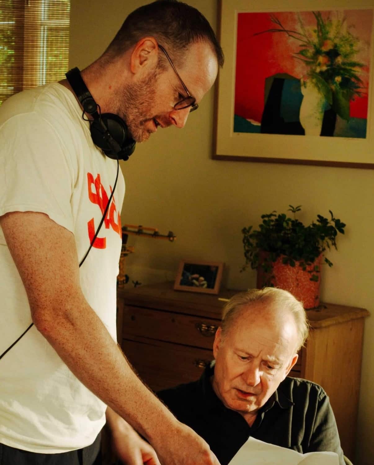 A man wearing headphones leans over an older man to review a document together in a room with warm lighting and a wooden dresser.