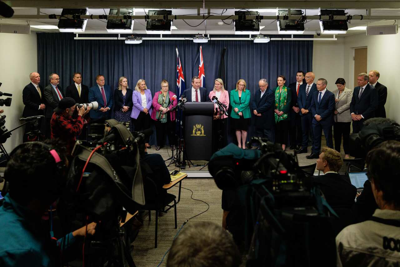 People flank a man standing at a lectern speaking in front of microphones.