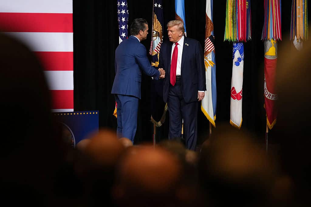 Two man in suits shake hands on stage in front of some flags.