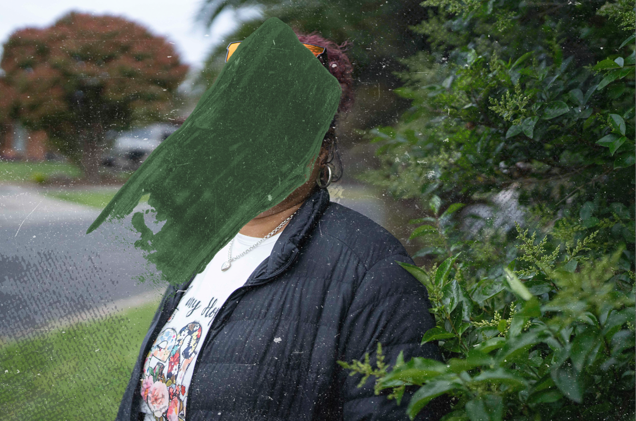 A woman wearing a black puffer jacket and a white t-shirt with colourful images on it stands outside near bushes. Her face is obscured by a black smudge.