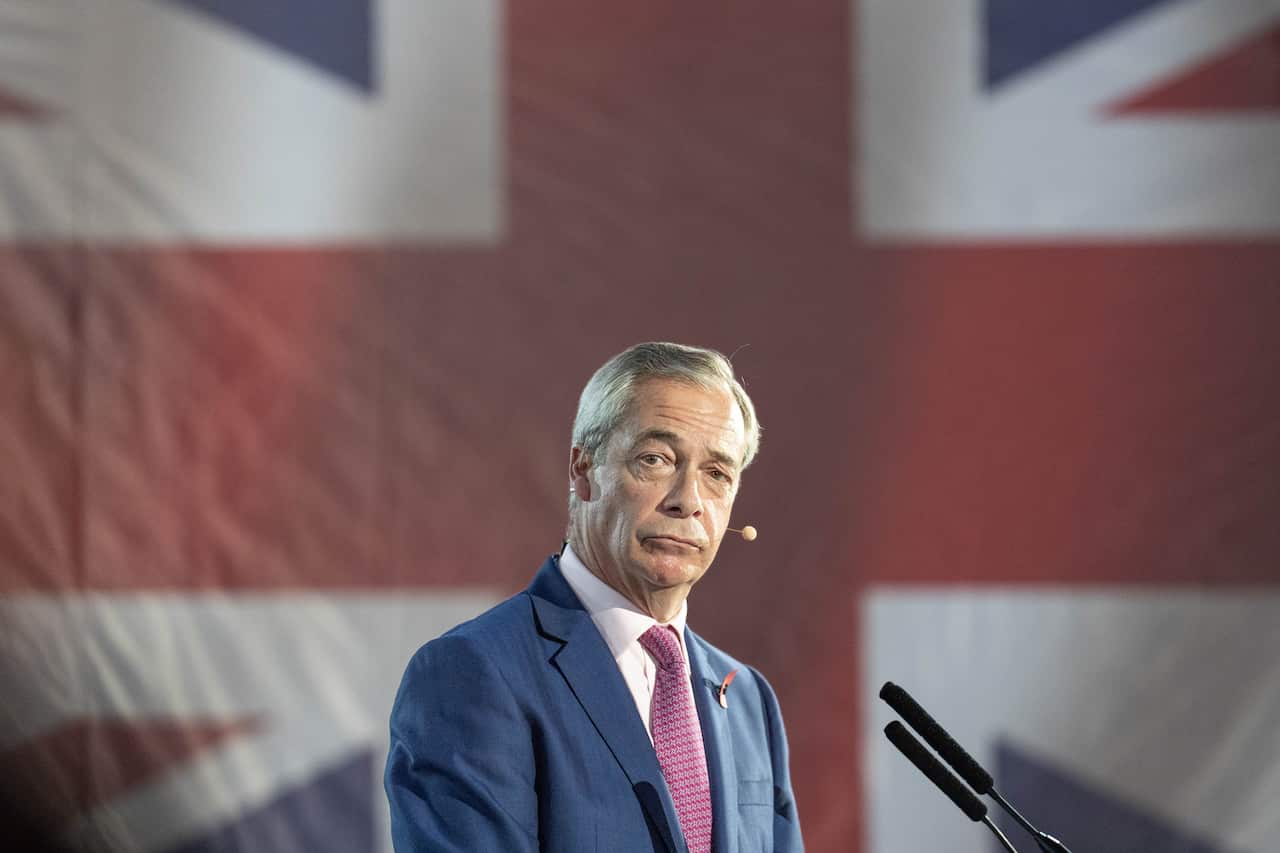 A man wearing a blue blazer, white shirt and red tie, standing in front of a giant British flag.