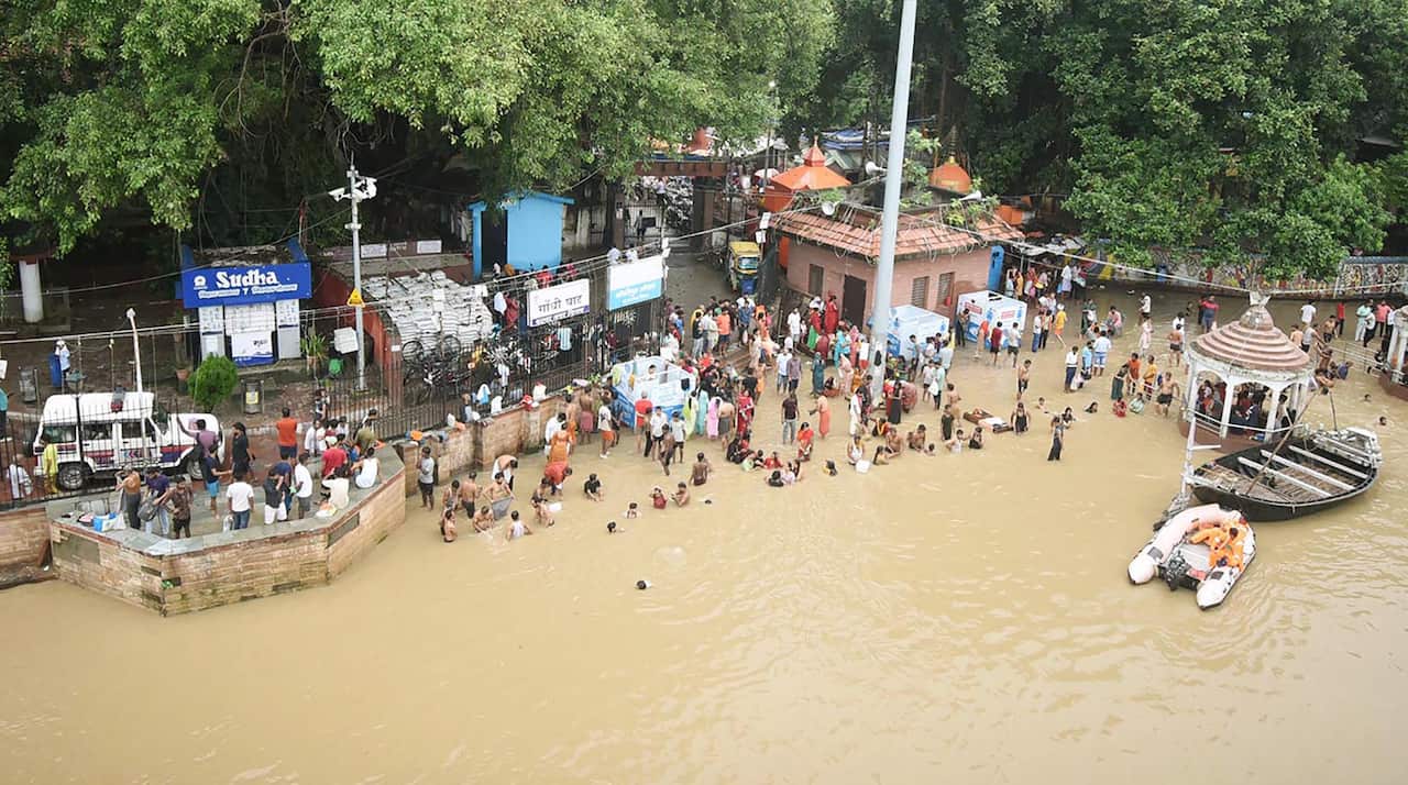 People gather and bathe in floodwaters at a riverside ghat surrounded by trees and partially submerged structures.