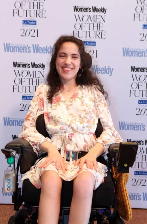 Hannah sits in a wheelchair in a floral dress behind a Women's Weekly backdrop. 