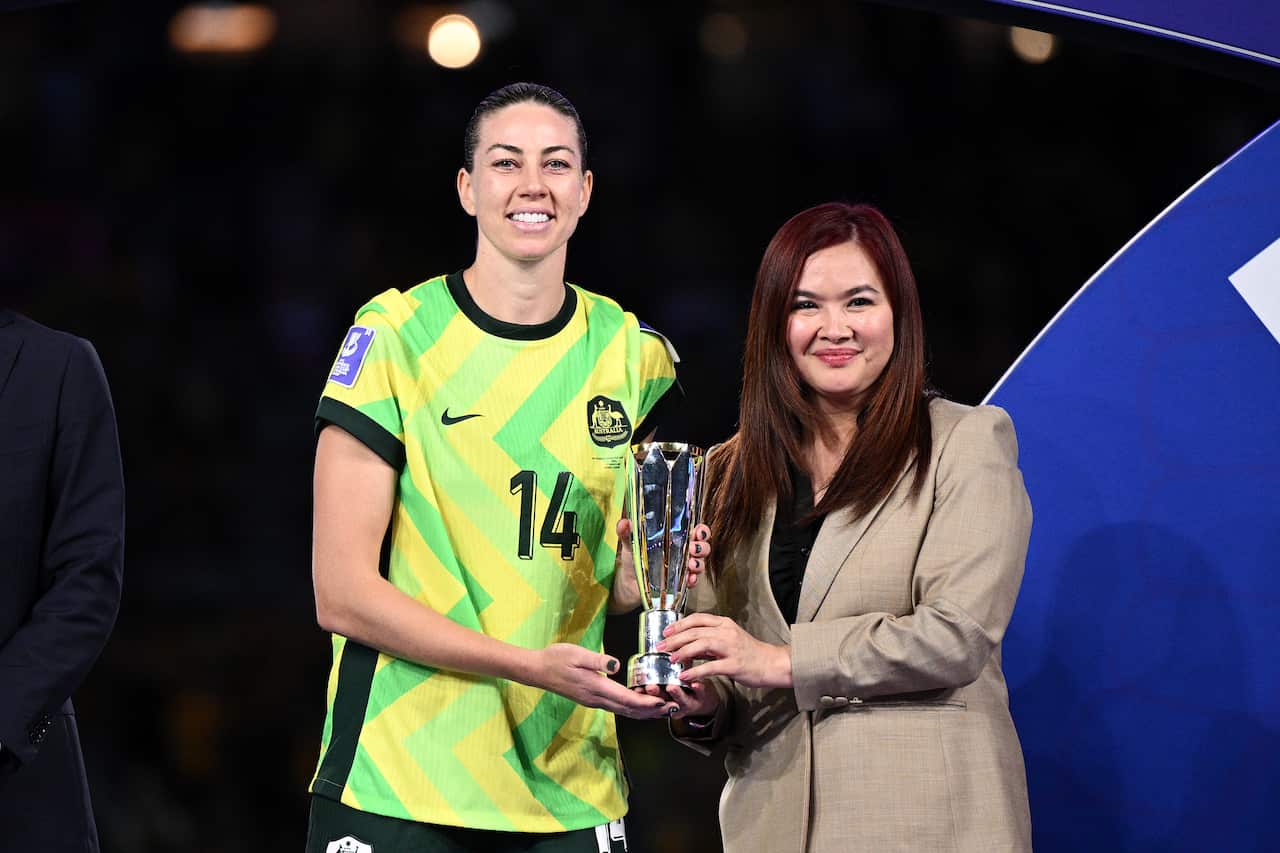 Matildas veteran Alanna Kennedy accepts the most valuable player of the tournament award following the 2026 Women's Asian Cup final. She is dressed in her green and yellow team kit holding a trophy alongside a match official.