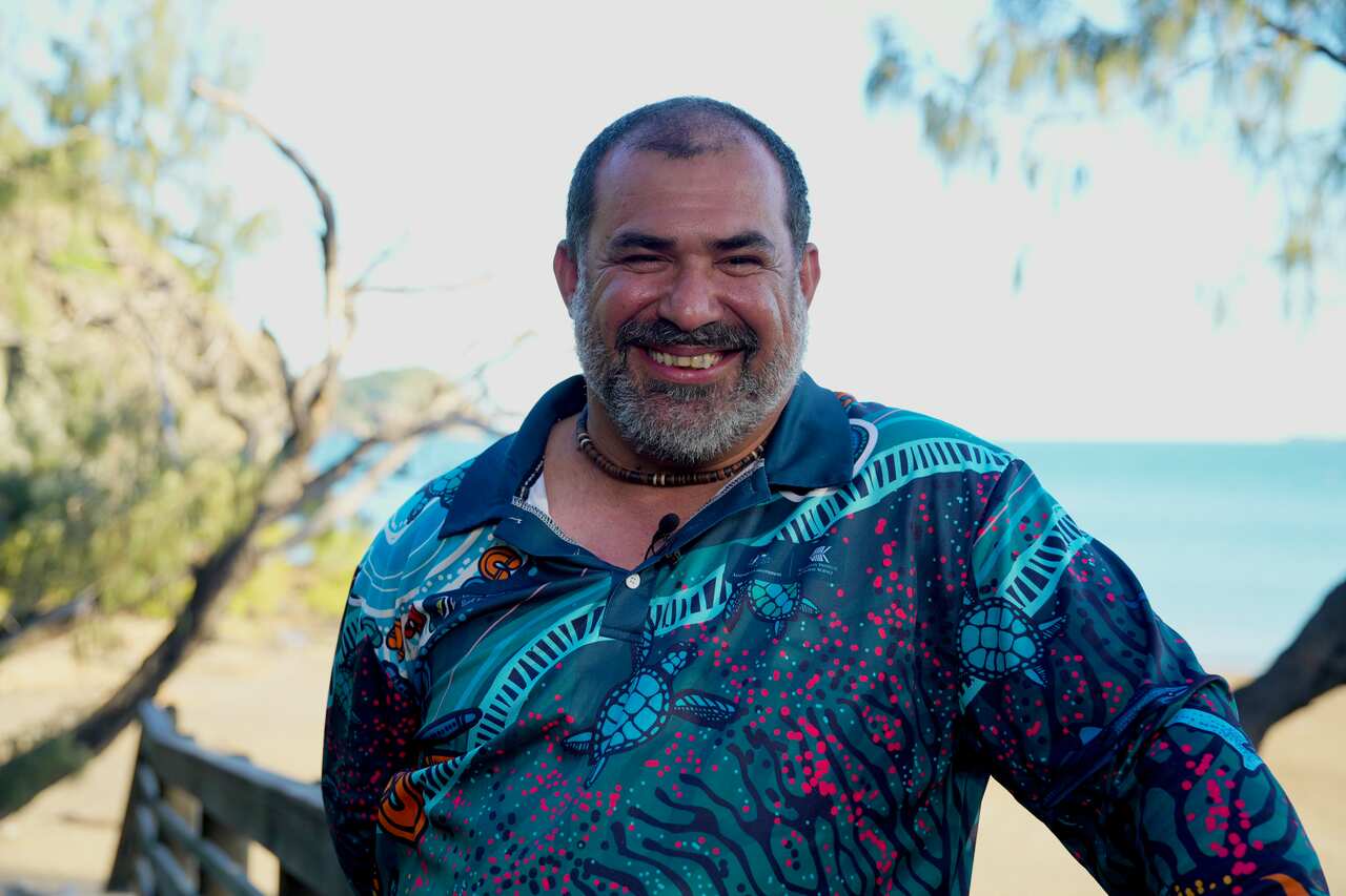 Man smiling wearing blue long sleeve shirt with beach behind him
