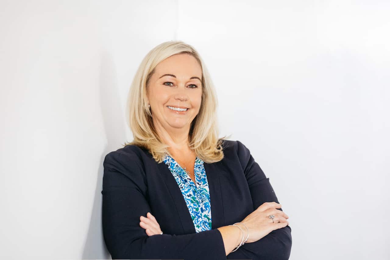 A woman in a blue shirt and dark blazer smiles to camera with her arms crossed