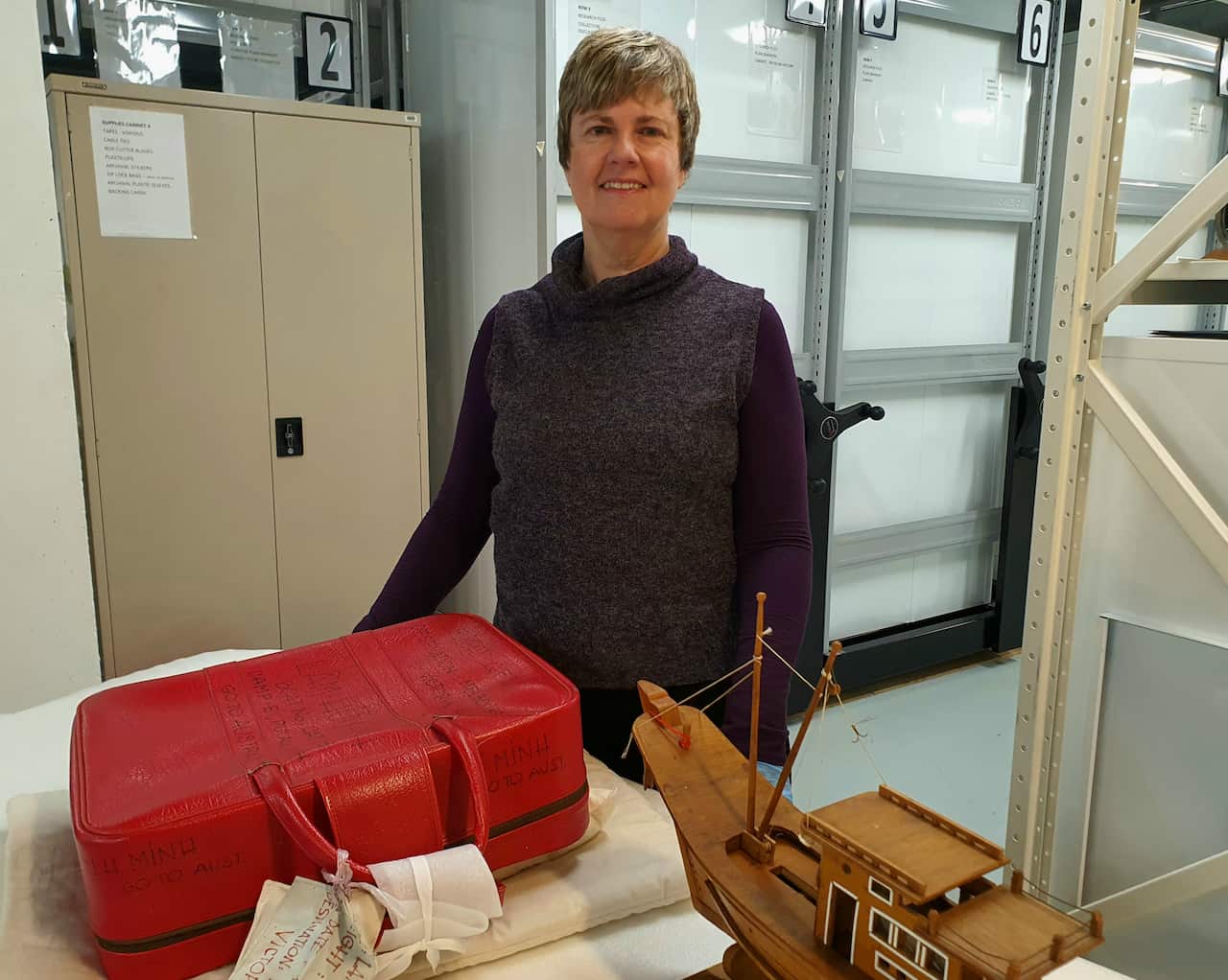 A middle aged woman standing in front of a red suitcase and wooden boat in a museum collections storeroom. 