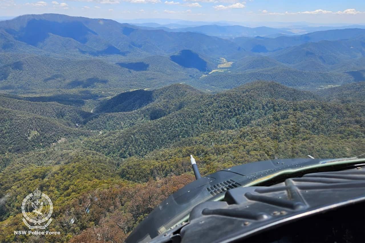 An image taken from the cockpit of a helicopter shows an aerial view of mountainous terrain covered in trees. 