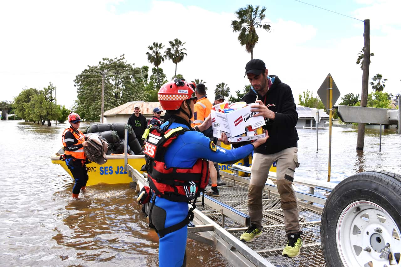 A man on a jetty hands a box of items to a rescue worker who is standing in floodwater.