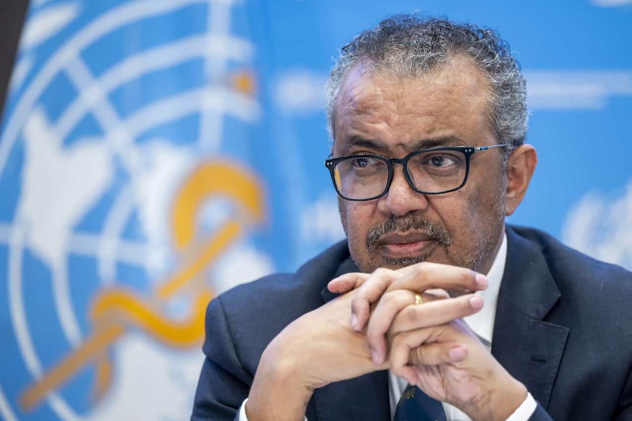 A man wearing glasses and a blue coat is sitting with his hands clasped together in front of a background with the United Nations logo.