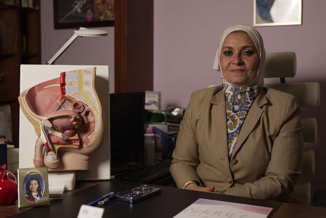  a woman in a cream hijab, a blue, white, and khaki patterned top, and a buttoned khaki blazer. She is sitting behind a desk against a pale pink wall and has a neutral facial expression. Next to her is a 3D diagram of the male reproductive system. On her desk is a document, a phone, a pen, and a small framed image of a young woman. 