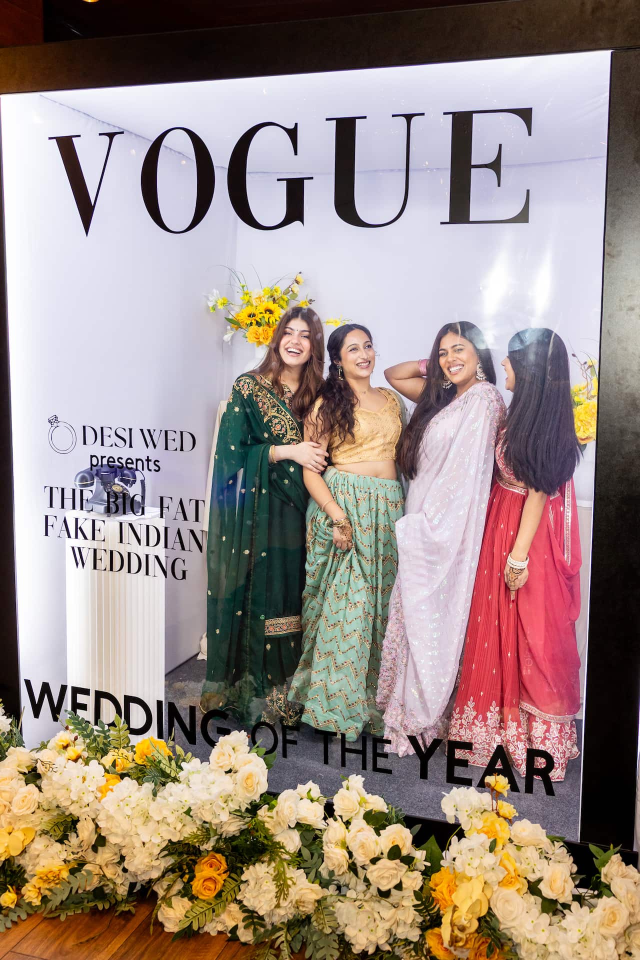 Four women in Indian wedding attire are posing together inside a large glass photo frame designed to look like a Vogue magazine cover.