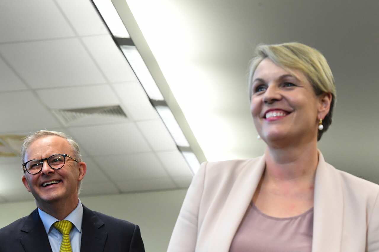 A white man with glasses laughs in the background as a white woman with blonde hair smiles