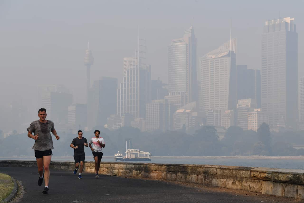 Three men run on a track near a harbour, with a smoky haze crossing over the water and tall buildings visible in the background.