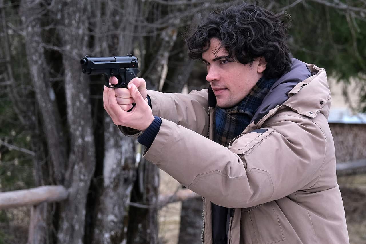 A young man with dark hair in a thick jacket and scarf holds a gun in two hands, pointed in front of him.