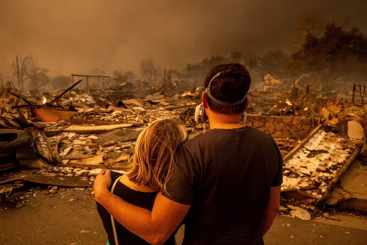A couple, standing and embracing each other with their backs to the camera, looks at the devastation caused by the fires.