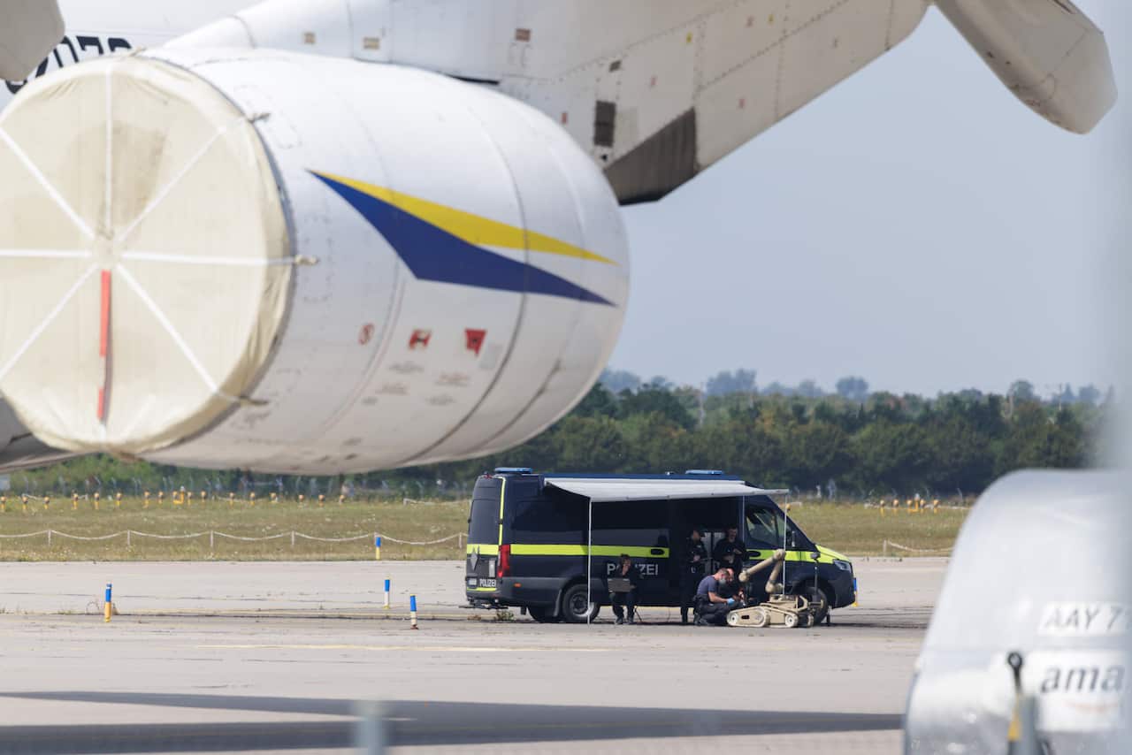 Police on an airport tarmac beside a van working on a robot designed to defuse bombs. The wing of a plane is in the foreground.