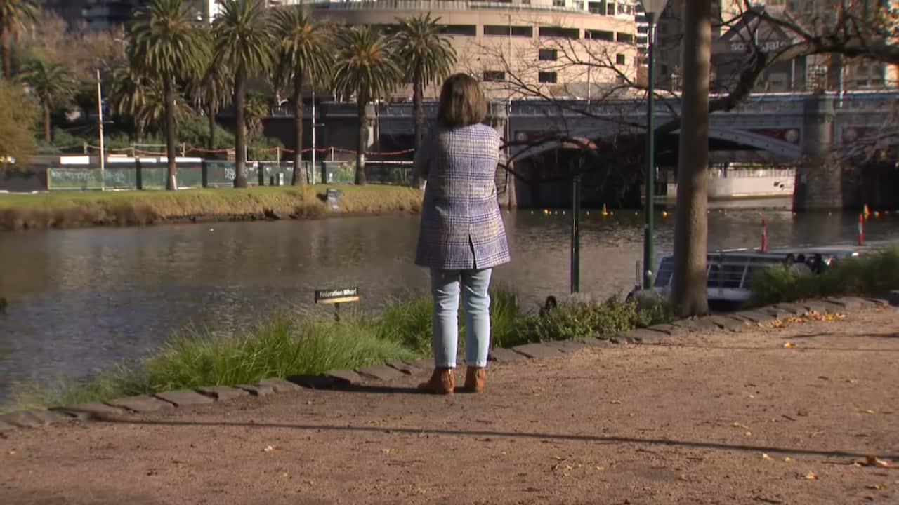 Woman in a checked coat standing with arms crossed on riverbank. 
