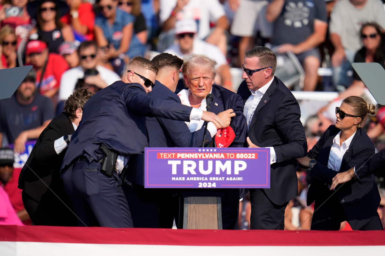 Donald Trump being shielded by Secret Service agents while at a podium. People in bleachers are behind him.