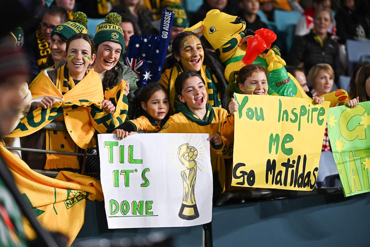 Girls wearing green and gold jerseys and holding up signs in a stadium in support of the Matildas 