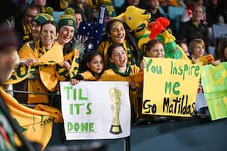 Girls wearing green and gold jerseys and holding up signs in a stadium in support of the Matildas