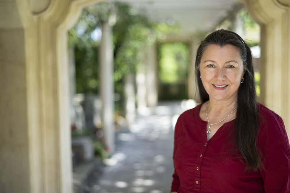 A woman in red top and long brown hair smiles at the camera. There is a shady verandah in the background.