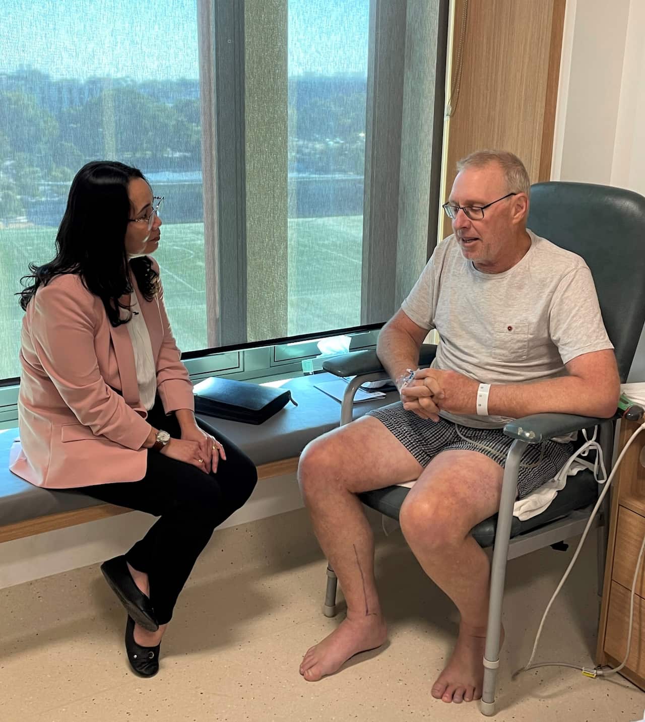 A woman in a pink jacket sits talking to a man in a hospital room.