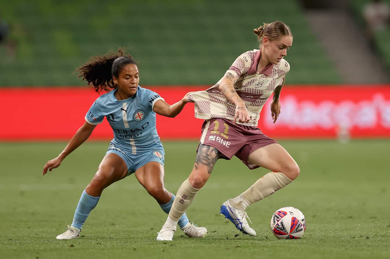 A female footballer in a blue kit grabs at the shirt of another female footballer wearing a cream and maroon jersey who is trying to kick the ball.
