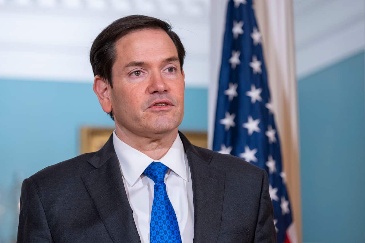 A man in a black blazer, white shirt and blue tie, speaking in front of an American flag.