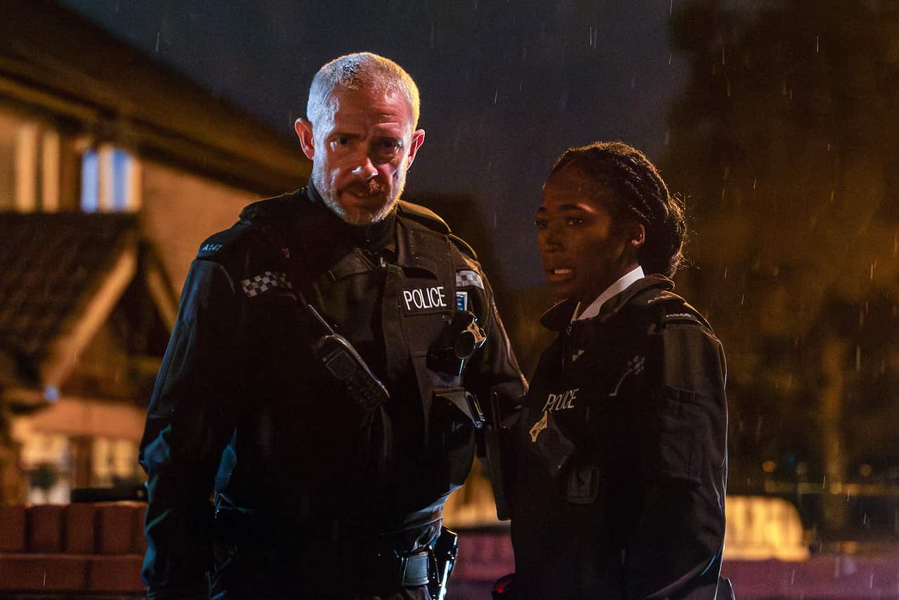 A policeman and policewoman stand in a street at night. It is raining and they both look worried.