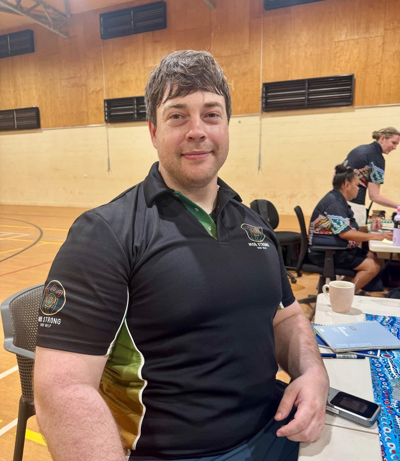 Dunghutti man Mark Holden sitting at a trestle table with his laptop smiling at the camera wearing a Strong Mob Debt Help polo t-shirt