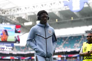 A black football player wearing headphones and a hoodie jumper with his arms in the pockets