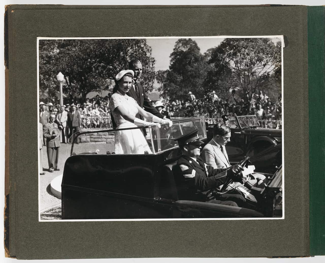 Old, grey photo of a young Queen Elizabeth and the Duke of Edinburgh standing in the back of a car with a crowd of people in the background