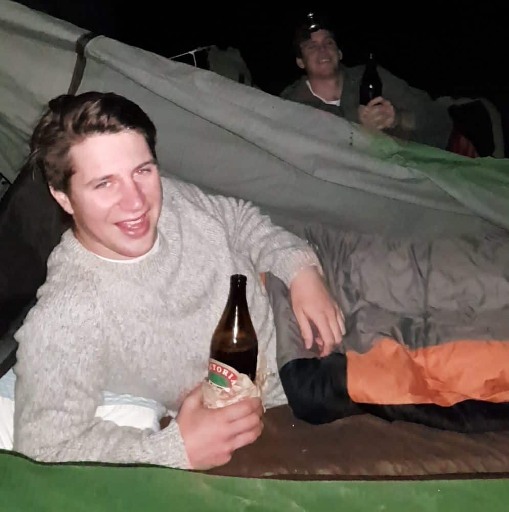 Young man in a sleeping bag holding a beer and smiling