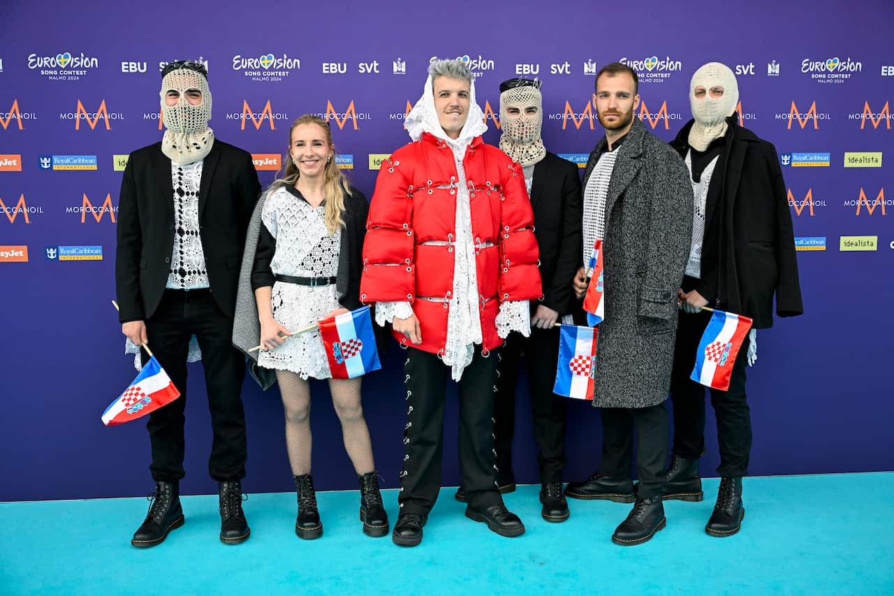 Six people in mostly black and white costumes stand in front of a purple wall with advertising on it