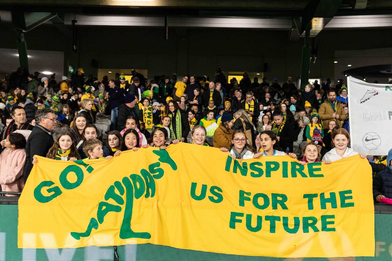 Fans at women's football match holding a yellow banner that reads: Go! Matildas inspire us for the future