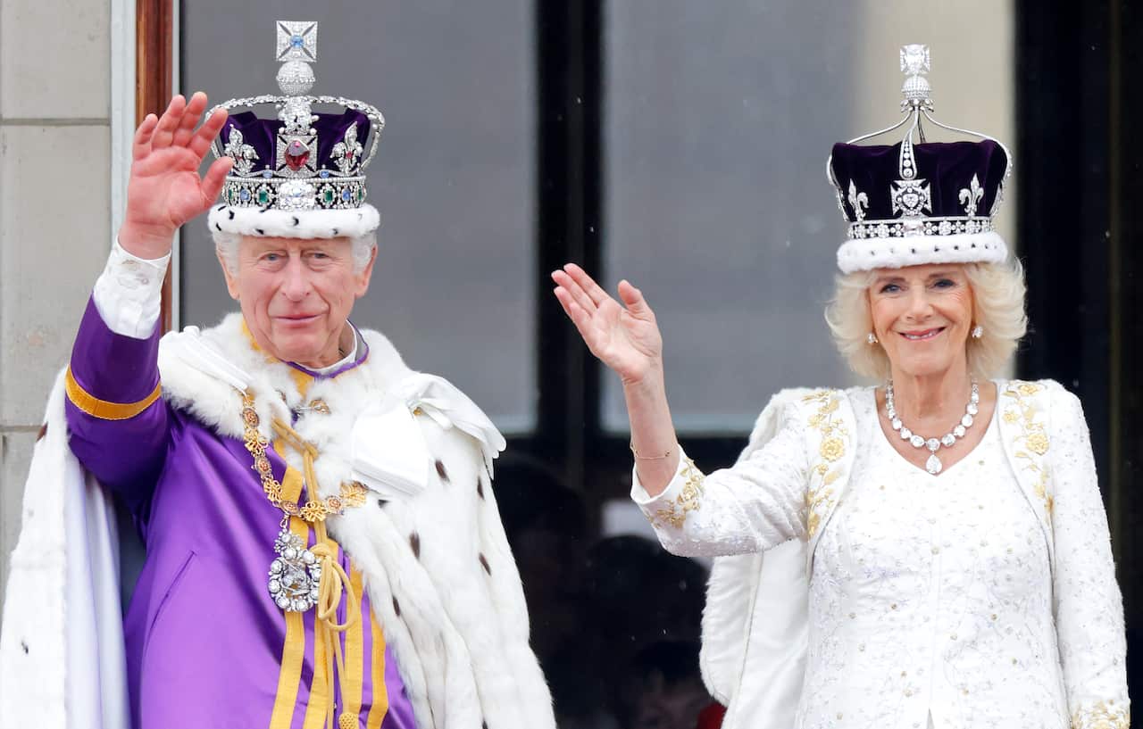 A man and woman, both in regal clothing, waving on a balcony.