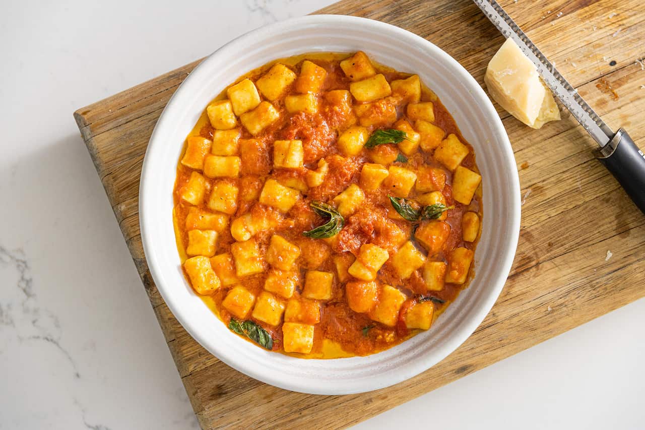 A white bowl sitting on a wooden board holds gnocchi in a tomato sauce. 