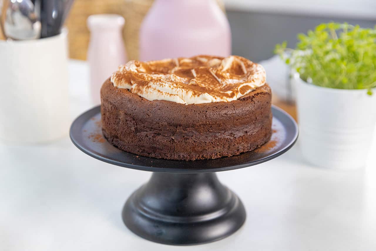 A deep chocolate cake, with a creamy topping, sits on a black cake stand.
