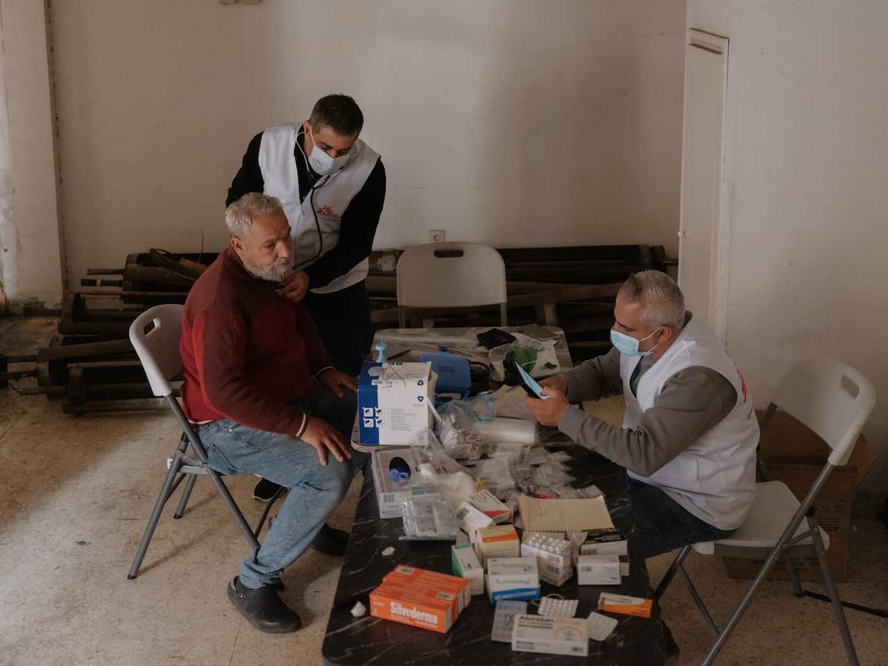 A doctor sits at a wooden table in a clinic with an elderly male patient across from him. A nurse is examining the patient as he sits.