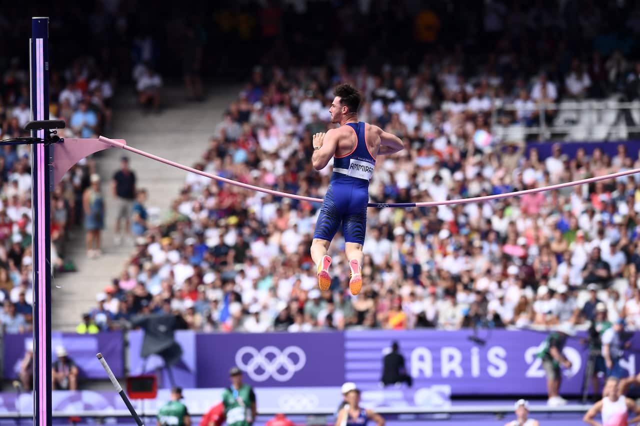 Rear view of a man in a blue athletics outfit contacting a horizontal bar high in the air.