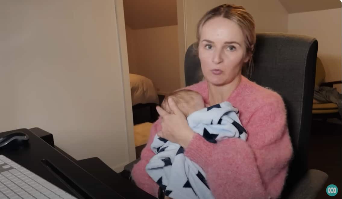 A blonde woman in a pink jumper holds a newborn to her chest while sitting in front of a computer.