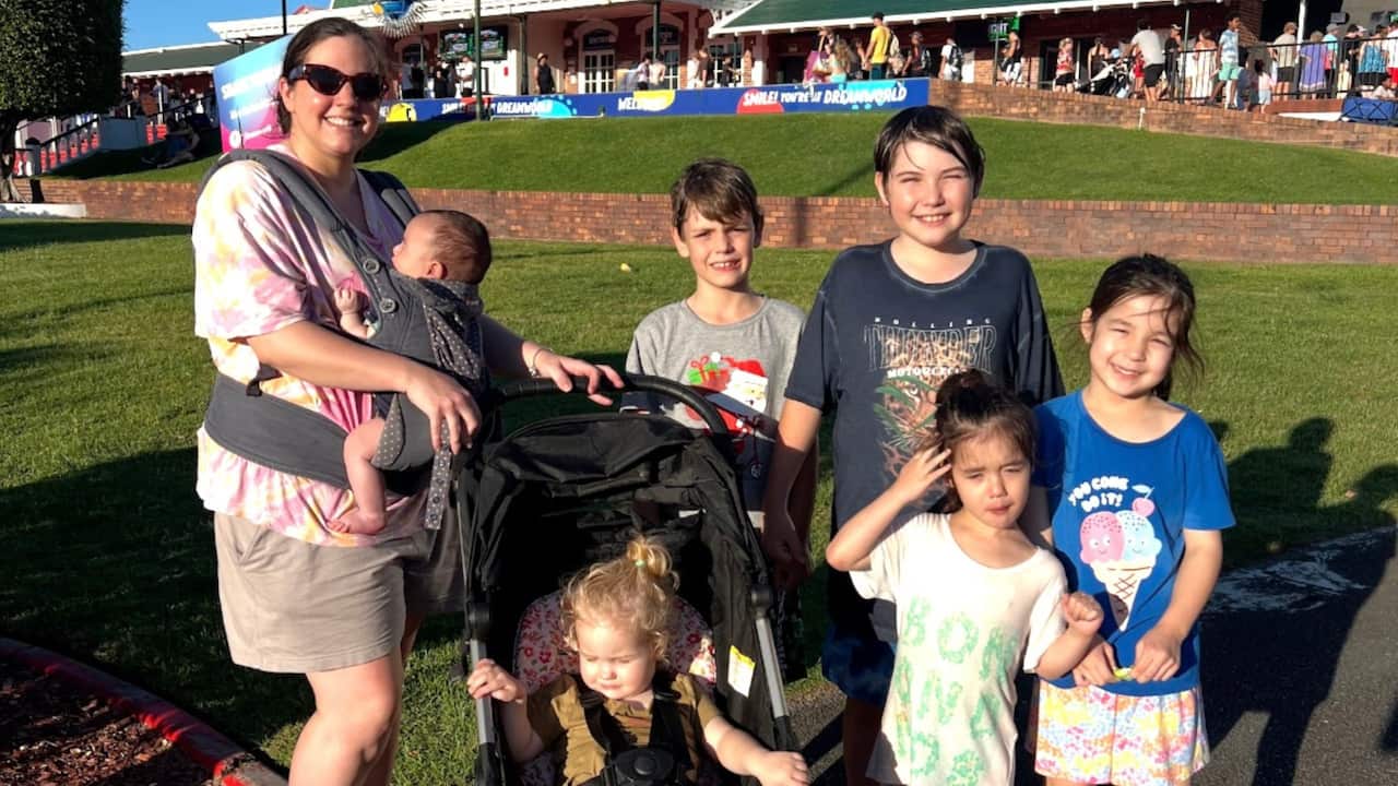 a young mum holding a baby in a carrier on her chest holds a pram with a toddler in it and four other young kids stand by the pram outside a theme park