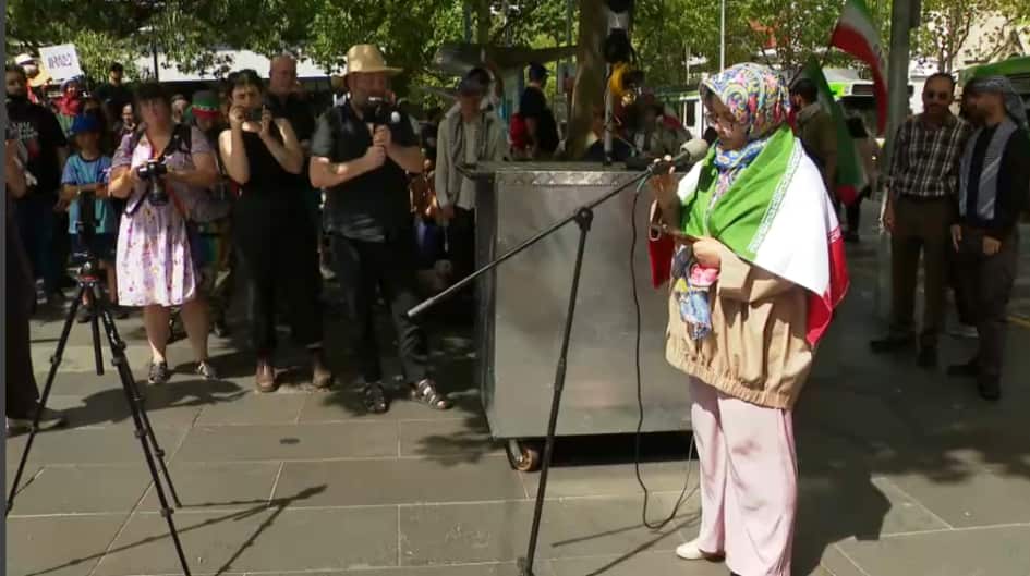 A woman draped in an Iranian flag and wearing a colorful headscarf speaks into a microphone at an outdoor gathering while several people look on and take photographs.