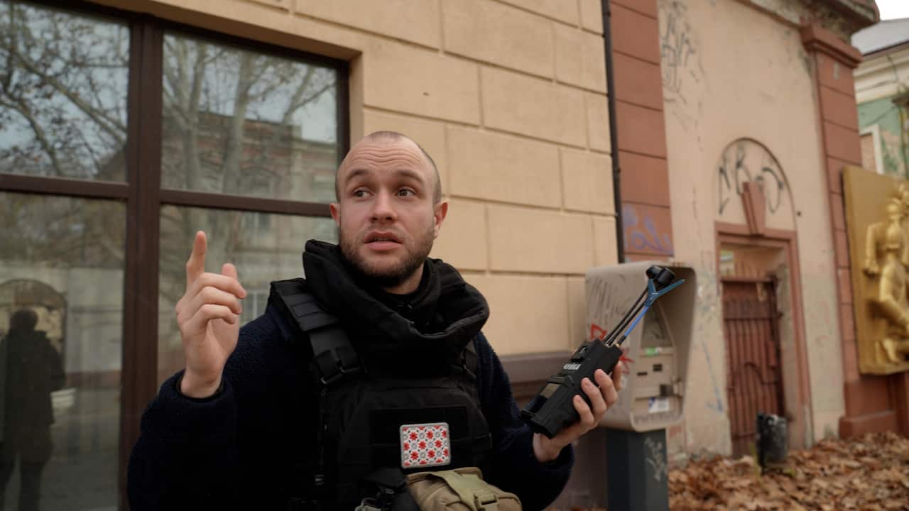 A man in a bulletproof vest holds a drone detector device as he looks about the street with a worried expression on his face.