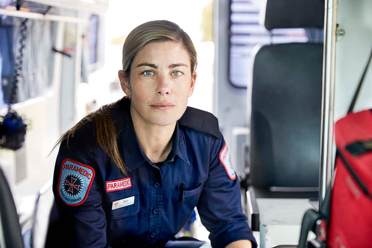 A woman in a paramedic uniform sits in an ambulance, looking forward with a calm face.