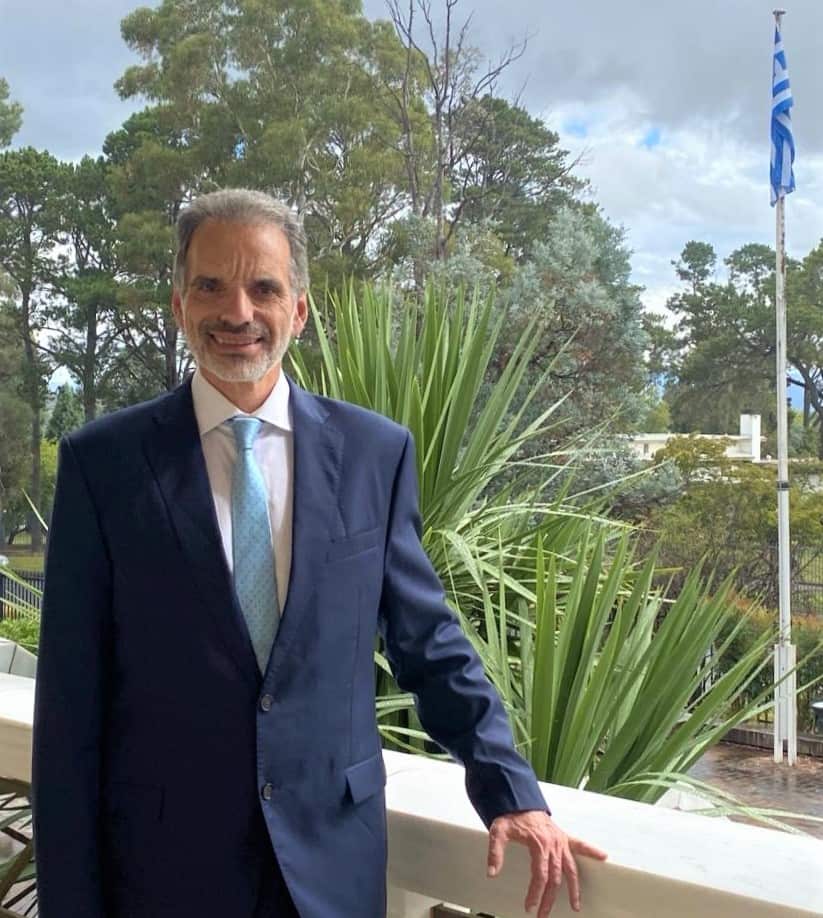 A man in a blue suit stands on a balcony with a Greek flag in the background.