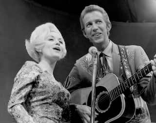 Black-and-white photo of a performer with a large hairstyle playing an acoustic guitar and singing into a microphone on stage.
