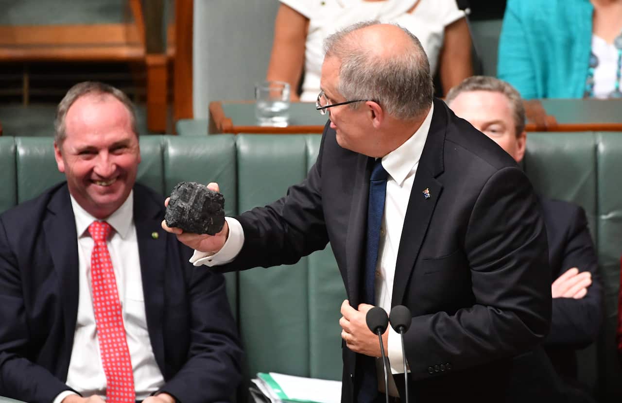 A man in a suit holds a lump of coal in parliament. Another quite red-faced man smiles behind him. 